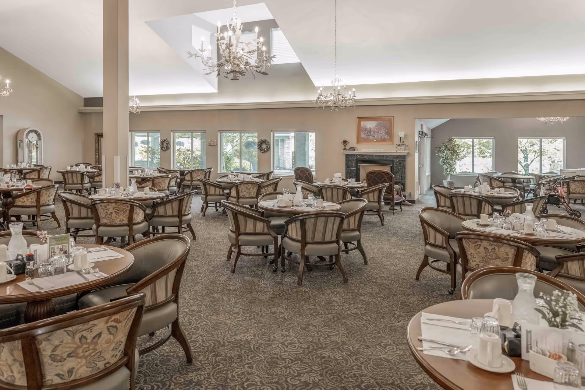 Spacious, elegant dining room with multiple round tables and upholstered chairs set for meals, chandeliers, and windows overlooking greenery.
