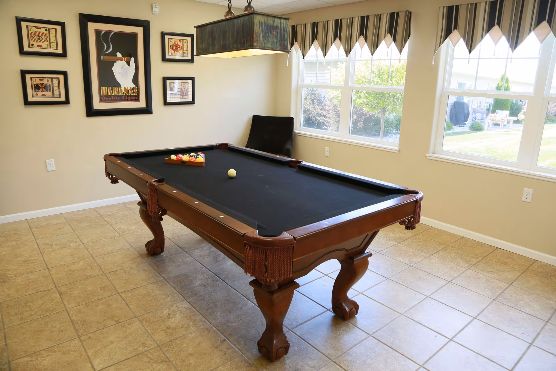 A room with a wooden pool table with a black felt surface, a triangle rack with billiard balls, and a cue ball. The room has beige tiled flooring, cream-colored walls, and large windows with striped valances letting in natural light. There are framed pictures on the wall, including one with a hand holding a cigar and the word 'Habanas'. A black chair is placed near the windows.