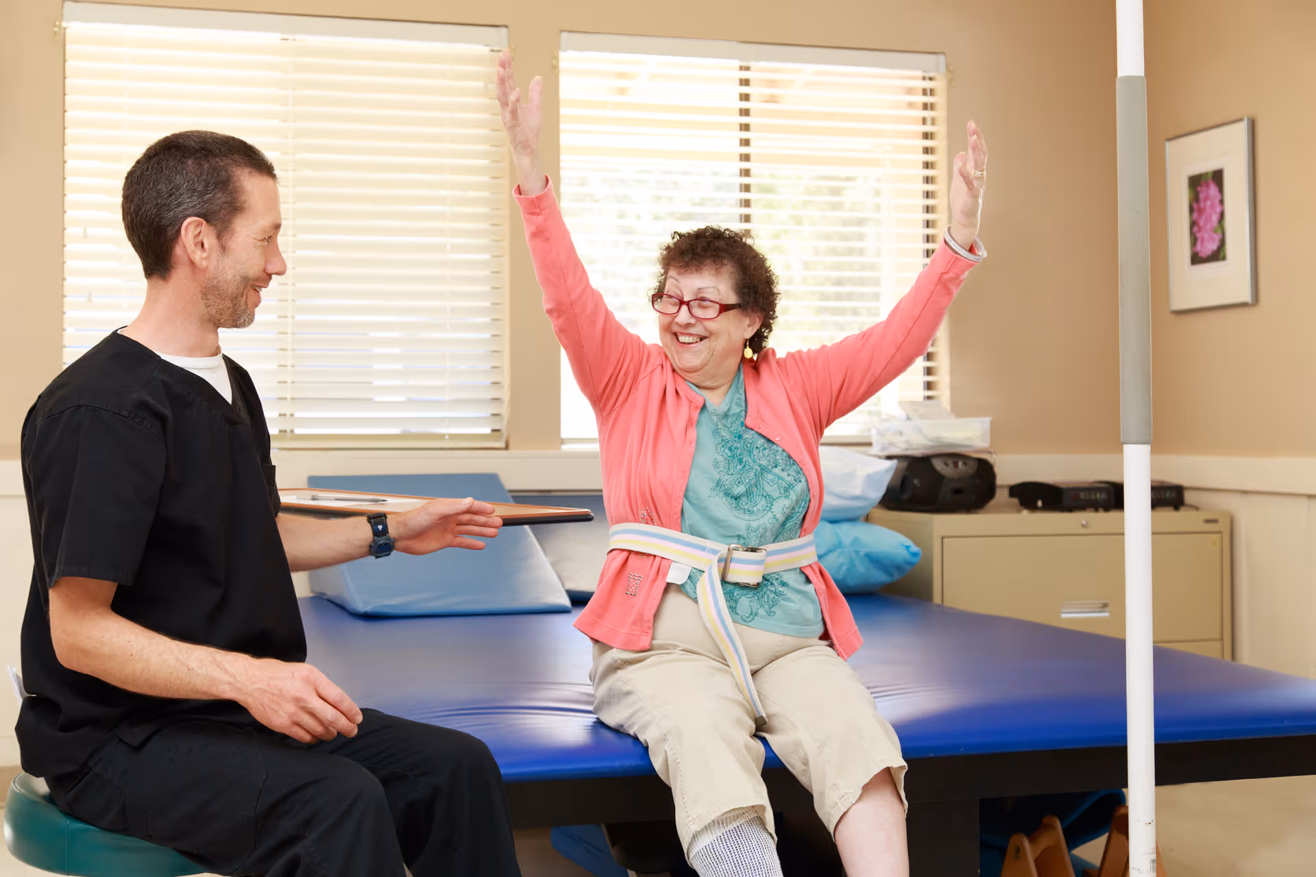 A senior woman wearing a pink cardigan and beige pants is sitting on a blue therapy table with her arms raised, smiling. A male healthcare professional in black scrubs is sitting nearby, engaging with her in a bright room with windows and blinds.