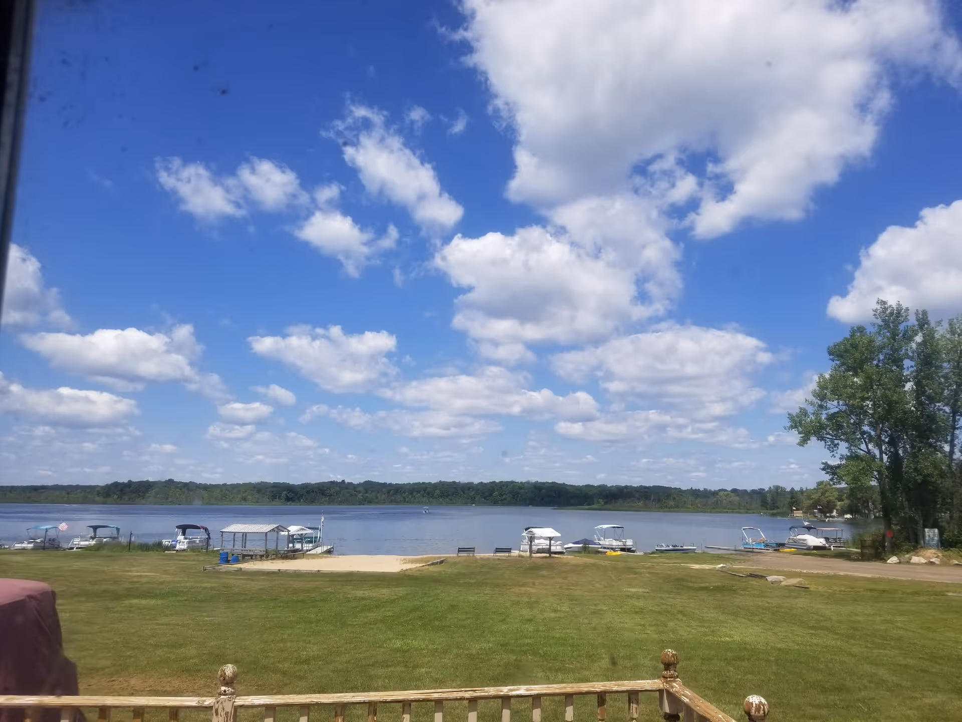 View of a calm lake with several boats docked along the shore, a grassy area in the foreground, and a partly cloudy blue sky above.