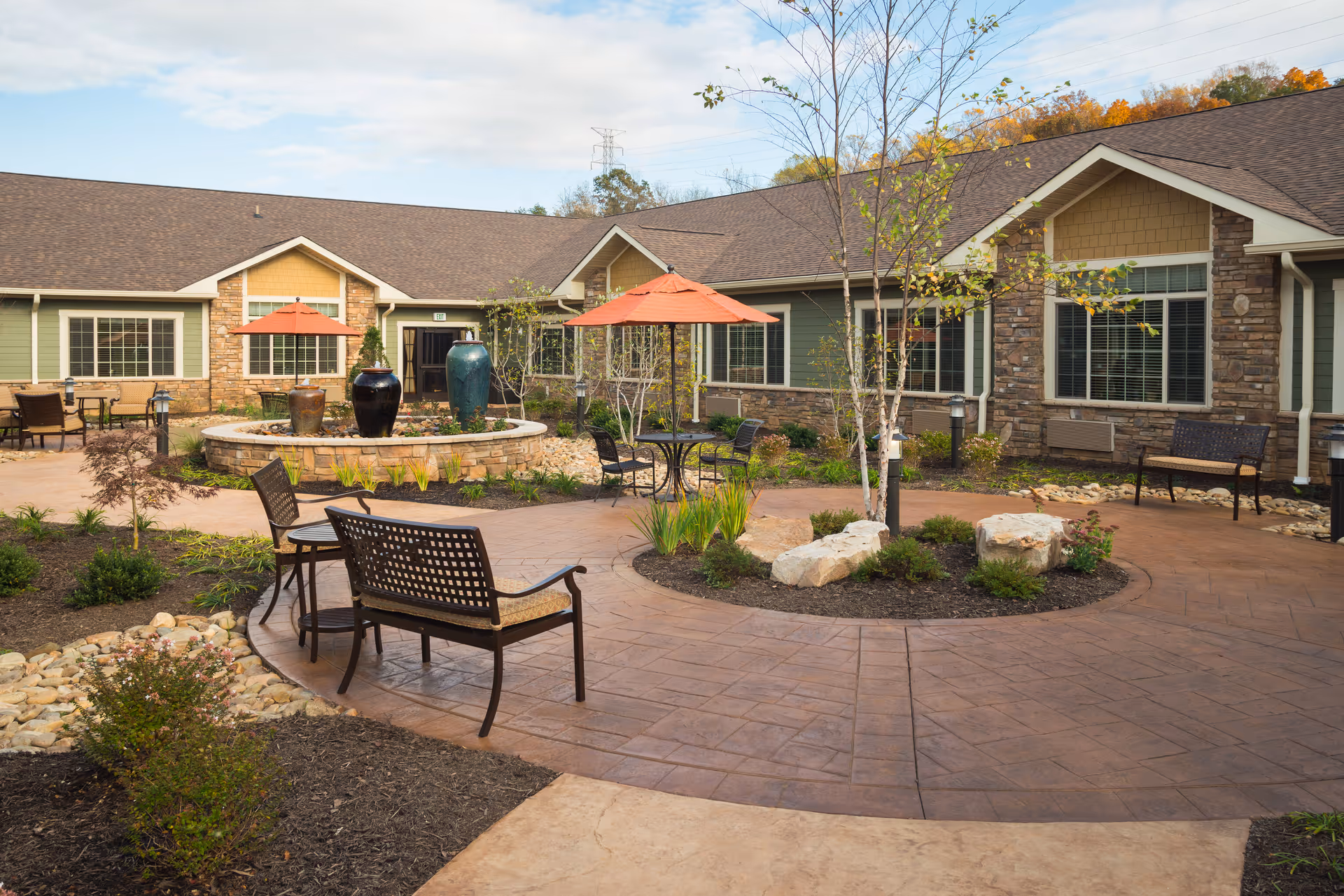 Outdoor courtyard area of a senior living facility with paved walkways, benches, tables with orange umbrellas, decorative planters, and landscaped greenery surrounded by a single-story building with green and beige siding and stone accents.