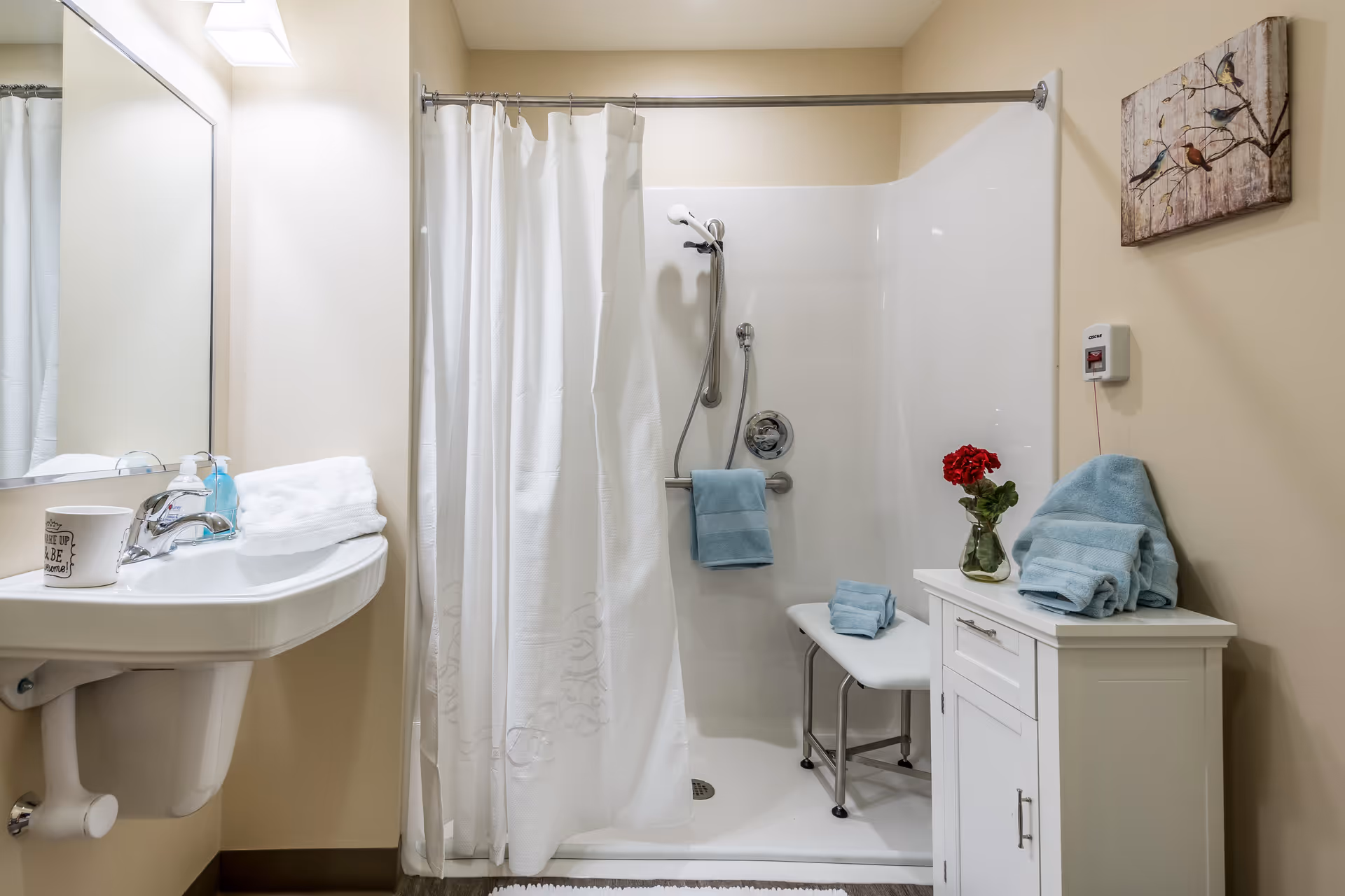 A clean and accessible bathroom featuring a walk-in shower with a white curtain, a shower seat, and grab bars. There is a white sink with a mirror above it on the left side, and a small white cabinet on the right side with folded blue towels and a vase with red flowers. A piece of wall art depicting birds on branches hangs above the cabinet.