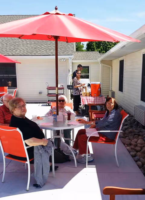 Four elderly people sitting around a white round table with red chairs under a large red patio umbrella outside a building. Two other people stand near a table covered with a red and white checkered tablecloth, which holds a large orange beverage dispenser and other items. The scene is bright and sunny with a clear blue sky.