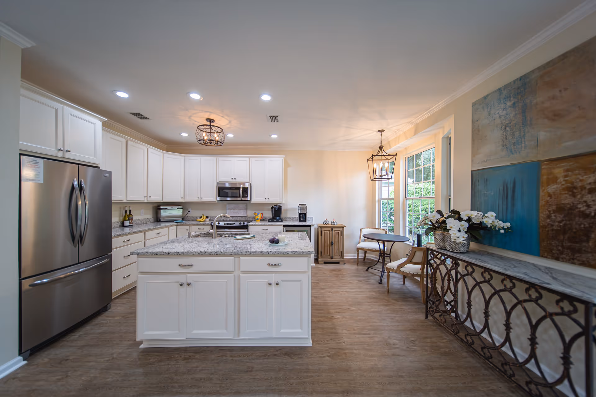 Bright and spacious kitchen with white cabinets, granite countertops, a large island in the center, stainless steel refrigerator, microwave, and stove. There is a small seating area with two chairs and a round table near large windows letting in natural light. Decorative paintings and a console table with flowers are along the right wall.