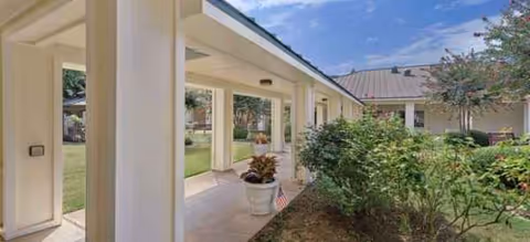 Exterior view of a covered walkway with white columns beside a landscaped garden.
