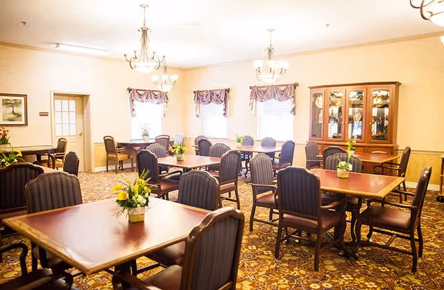 A bright and spacious dining room with multiple wooden tables and cushioned chairs arranged neatly. Each table has a small floral centerpiece. The room features patterned carpet, cream-colored walls, three windows with valances, and two chandeliers hanging from the ceiling. A wooden cabinet with glass doors displaying dishes is visible against one wall.