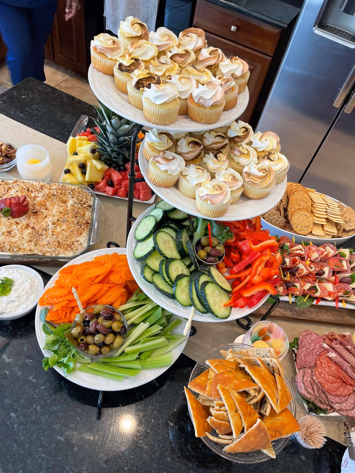 A kitchen counter displaying a variety of food items including a three-tiered stand with cupcakes on the top two tiers and sliced cucumbers, red bell peppers, and olives on the bottom tier. Surrounding the stand are plates with sliced carrots, celery, olives, crackers, assorted meats, pita bread, a fruit platter with pineapple and watermelon, a casserole dish, and a bowl of cottage cheese. A stainless steel refrigerator and wooden cabinets are visible in the background.