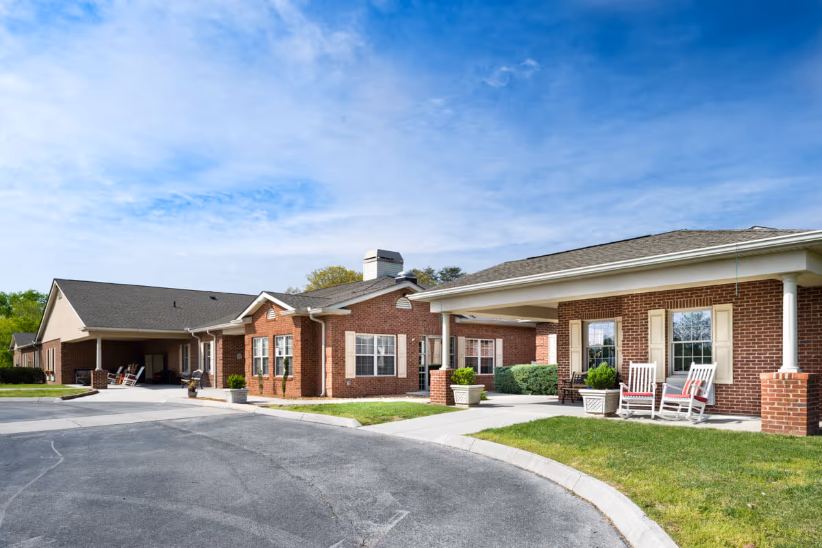 Front exterior of a single-story brick assisted living facility with a covered entrance, rocking chairs on the porch, and a driveway.