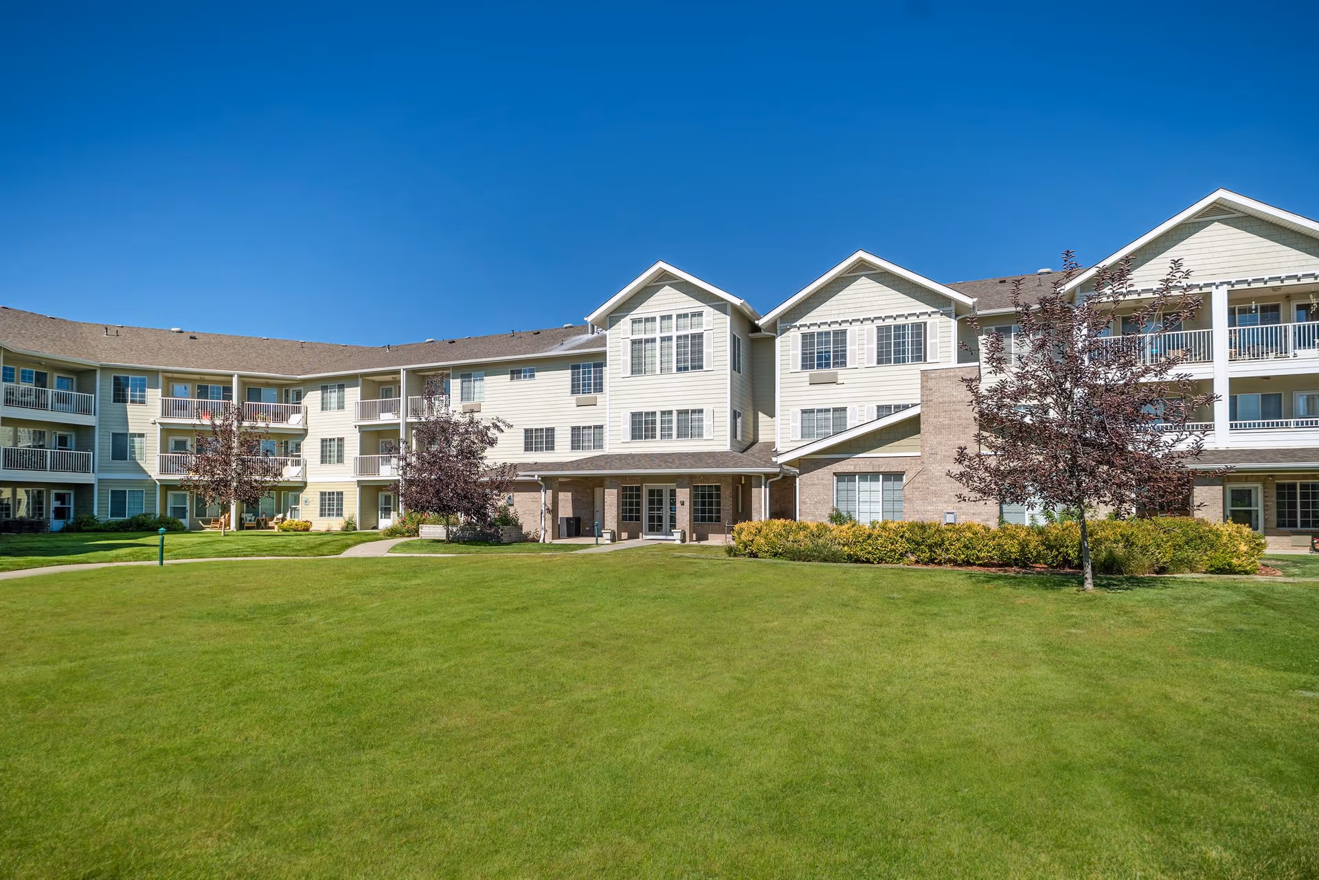 Exterior view of a three-story senior living facility building with beige siding and brick accents, surrounded by a well-maintained green lawn and a few small trees under a clear blue sky.