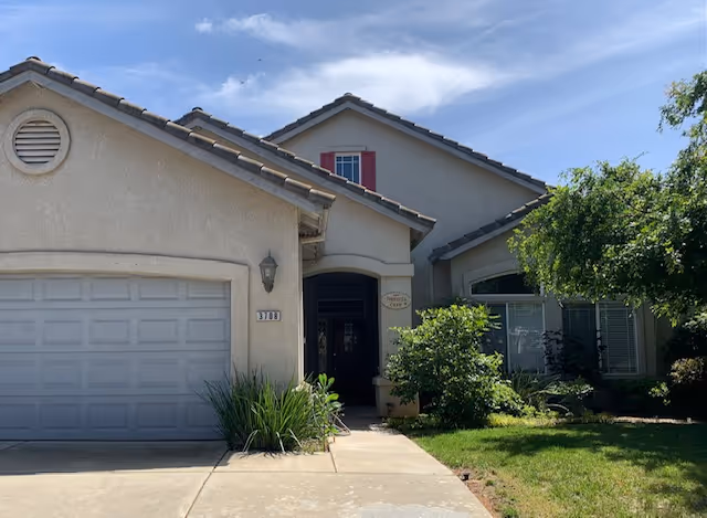 Exterior front view of a single-story residential-style building with a garage on the left, a driveway, and a small garden with bushes and a tree on the right under a partly cloudy sky.