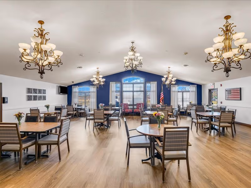 A spacious dining room with multiple round and square wooden tables surrounded by chairs. The room features large windows with white curtains, blue accent walls, and several ornate chandeliers hanging from the ceiling. There are flower vases on each table, an American flag near the windows, and framed artwork on the walls.