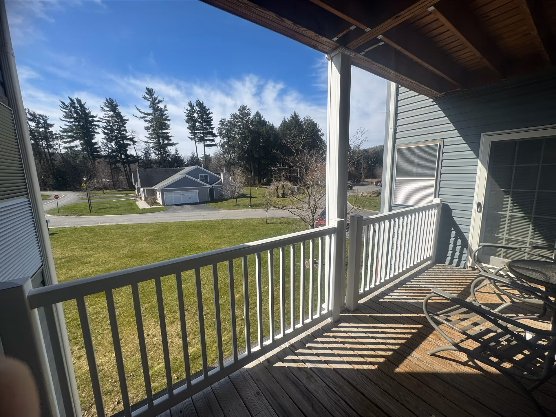 View from a covered balcony with wooden flooring and white railing overlooking a green lawn, a road, and a small building with garages. There are tall trees and a partly cloudy blue sky in the background. The balcony has a glass-top table and metal chairs.