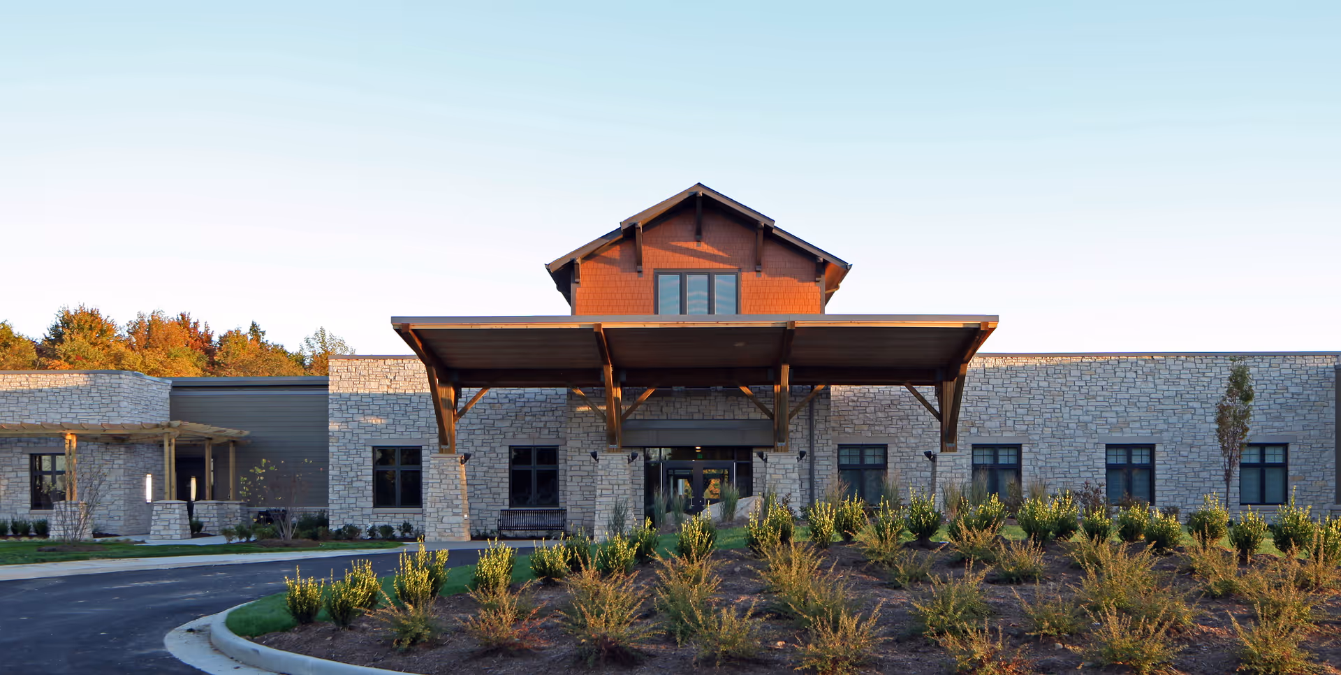 Front entrance of a single-story stone-faced building with a large wood-covered portico and landscaped beds in front.