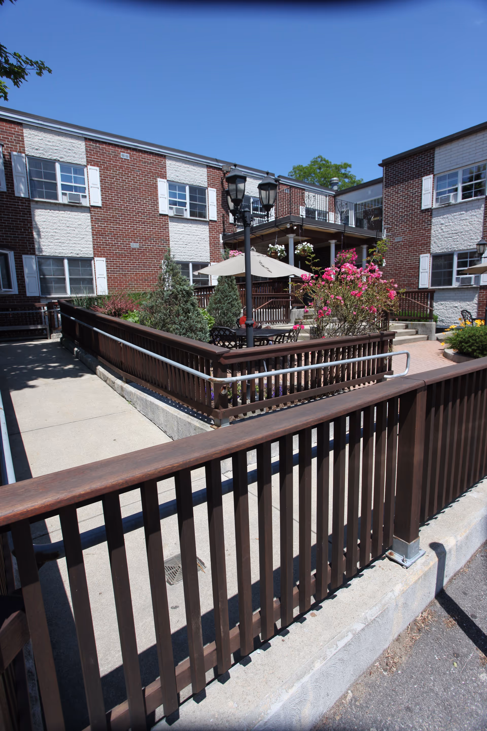Outdoor courtyard area of a senior living facility with brown wooden railings, concrete pathways, flowering bushes, patio tables with umbrellas, and a two-story brick building with white window shutters in the background under a clear blue sky.