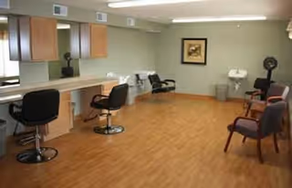 A spacious room with wooden flooring featuring salon-style chairs and mirrors along one wall, a sink, and additional seating including armchairs. The room is well-lit with overhead lighting and has a framed picture on the far wall.