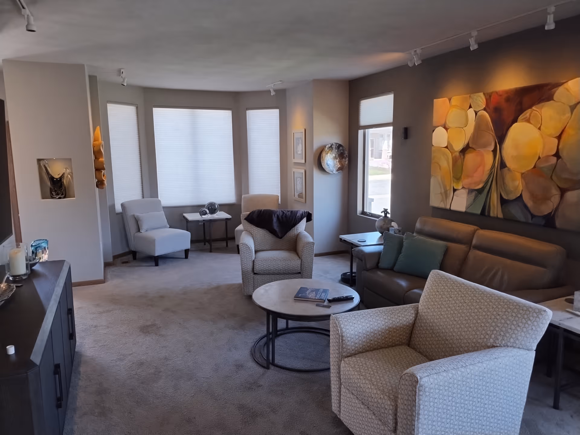 A cozy living room with beige carpet, a brown leather sofa with green pillows, two patterned armchairs, and a small round coffee table with a book and remote control. The room has large windows with white blinds, a side table with decorative items, and a colorful abstract painting on the wall.