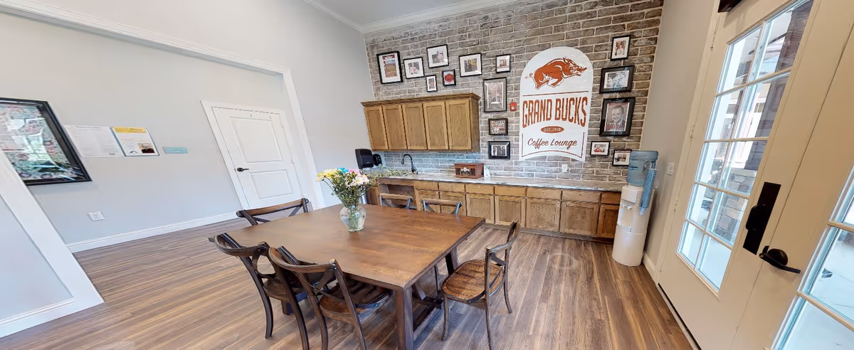 Communal coffee lounge with a wooden table and chairs, brick accent wall featuring a 'Grand Bucks Coffee Lounge' sign, cabinets and a water cooler.