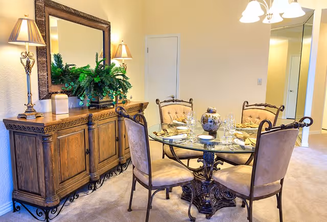 Elegant dining room with a glass-top round table set for four, upholstered chairs, and a wooden sideboard with lamps and a mirror.