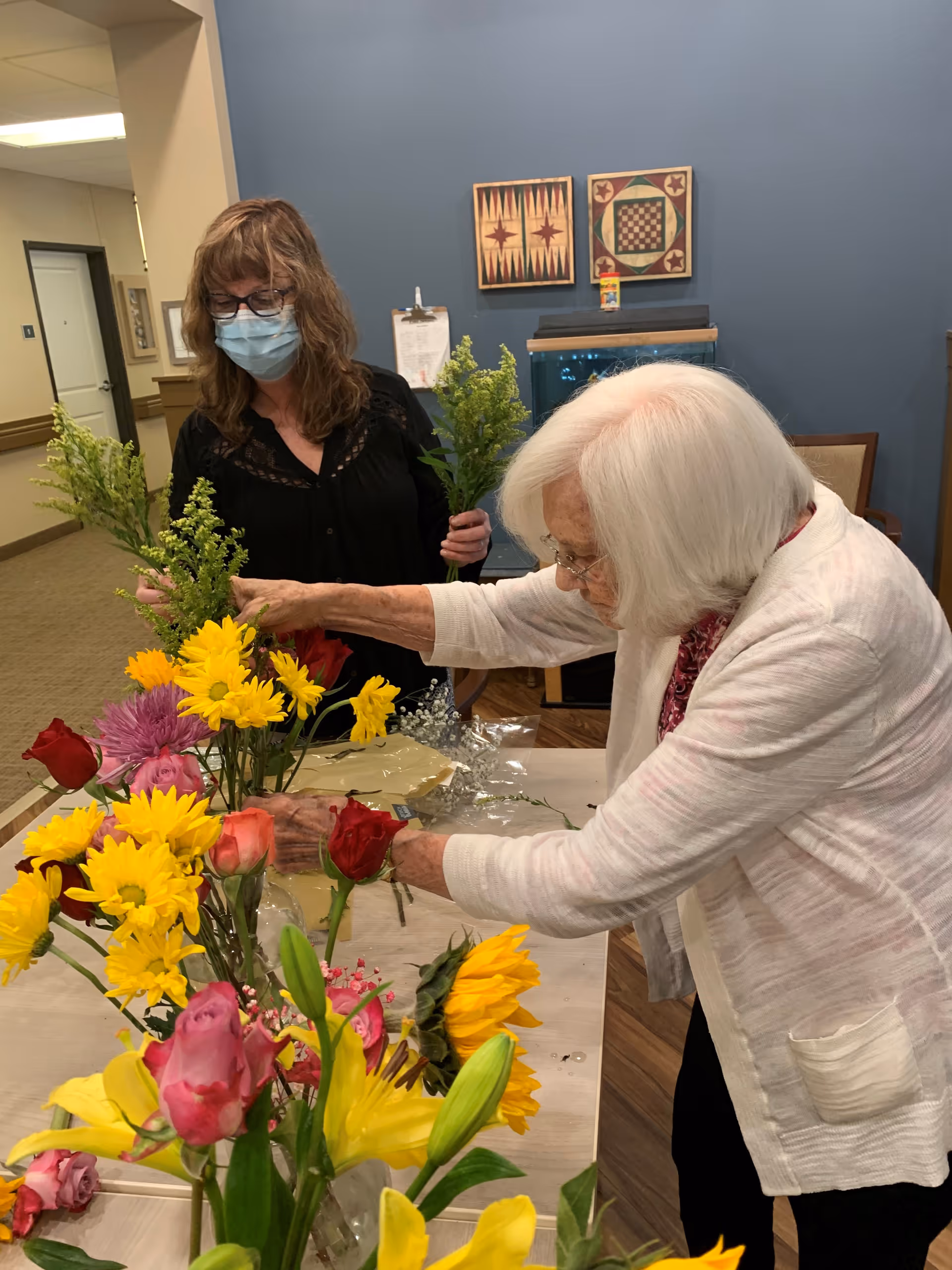 An elderly woman with white hair and glasses arranging flowers on a table filled with various colorful flowers, while a woman wearing a face mask and glasses stands nearby holding green foliage. They are in a room with blue walls decorated with framed artwork and a fish tank.