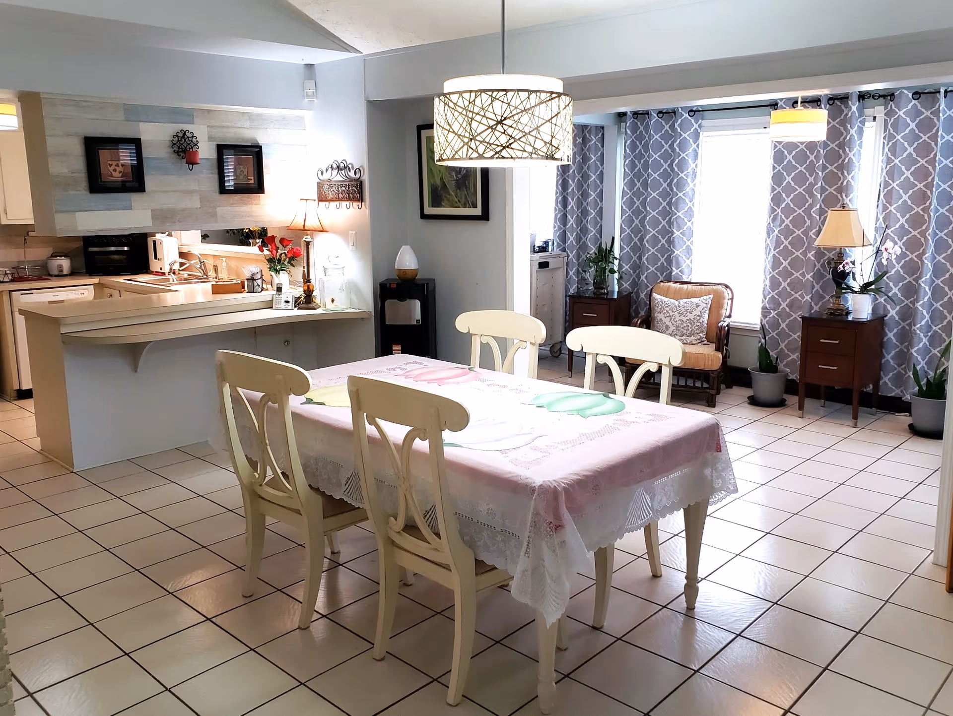Bright open dining area with a table and four chairs on a tiled floor, adjoining a kitchen and a sitting area with patterned curtains and lamps.