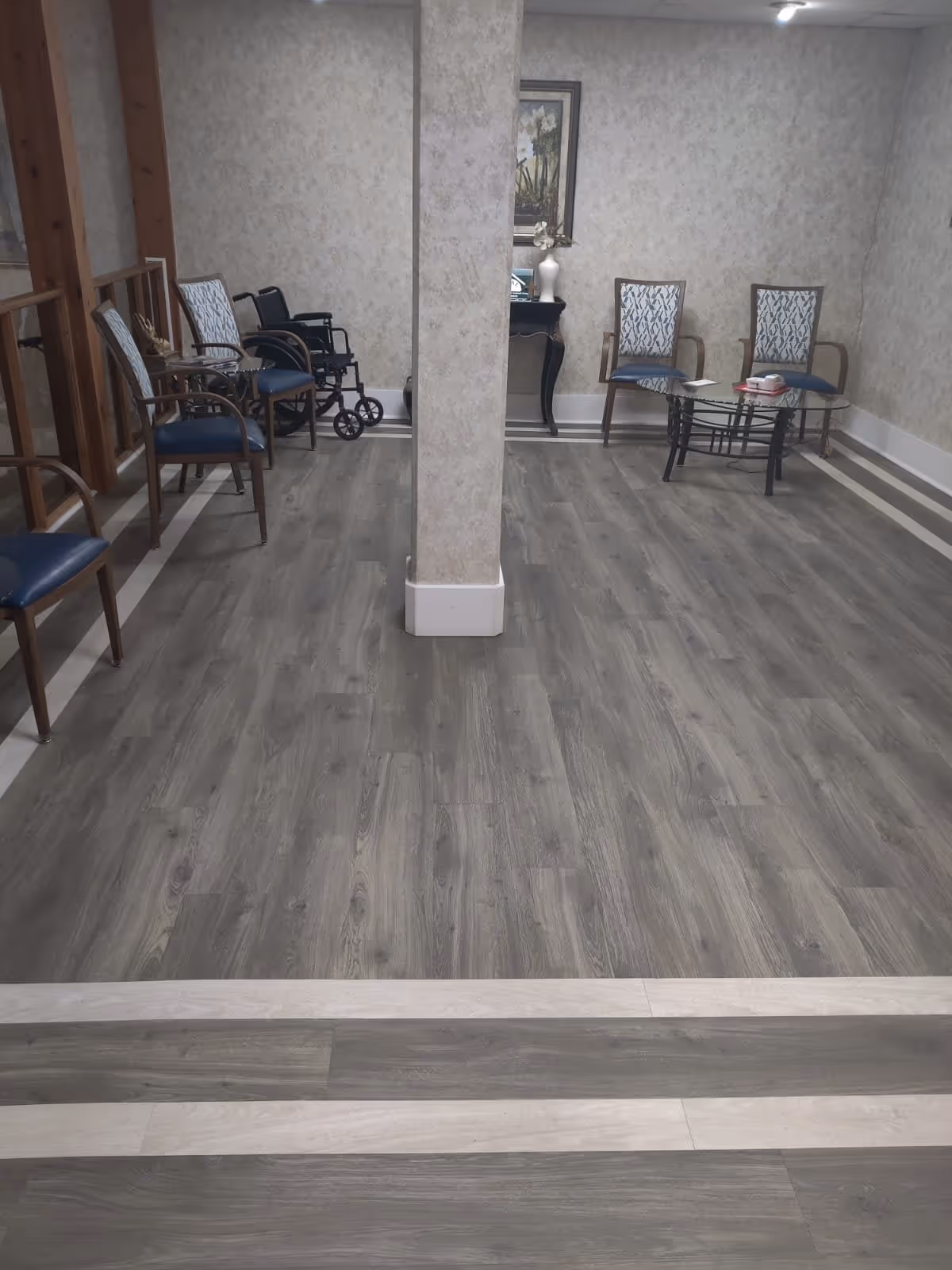A waiting area with several chairs arranged along the walls and a small glass coffee table in the center. The floor has wood-like vinyl planks with a light border. There is a wheelchair positioned near the chairs on the left side. A decorative table with a vase and framed artwork is visible against the back wall. The walls have a light textured wallpaper and a structural column stands in the middle of the room.