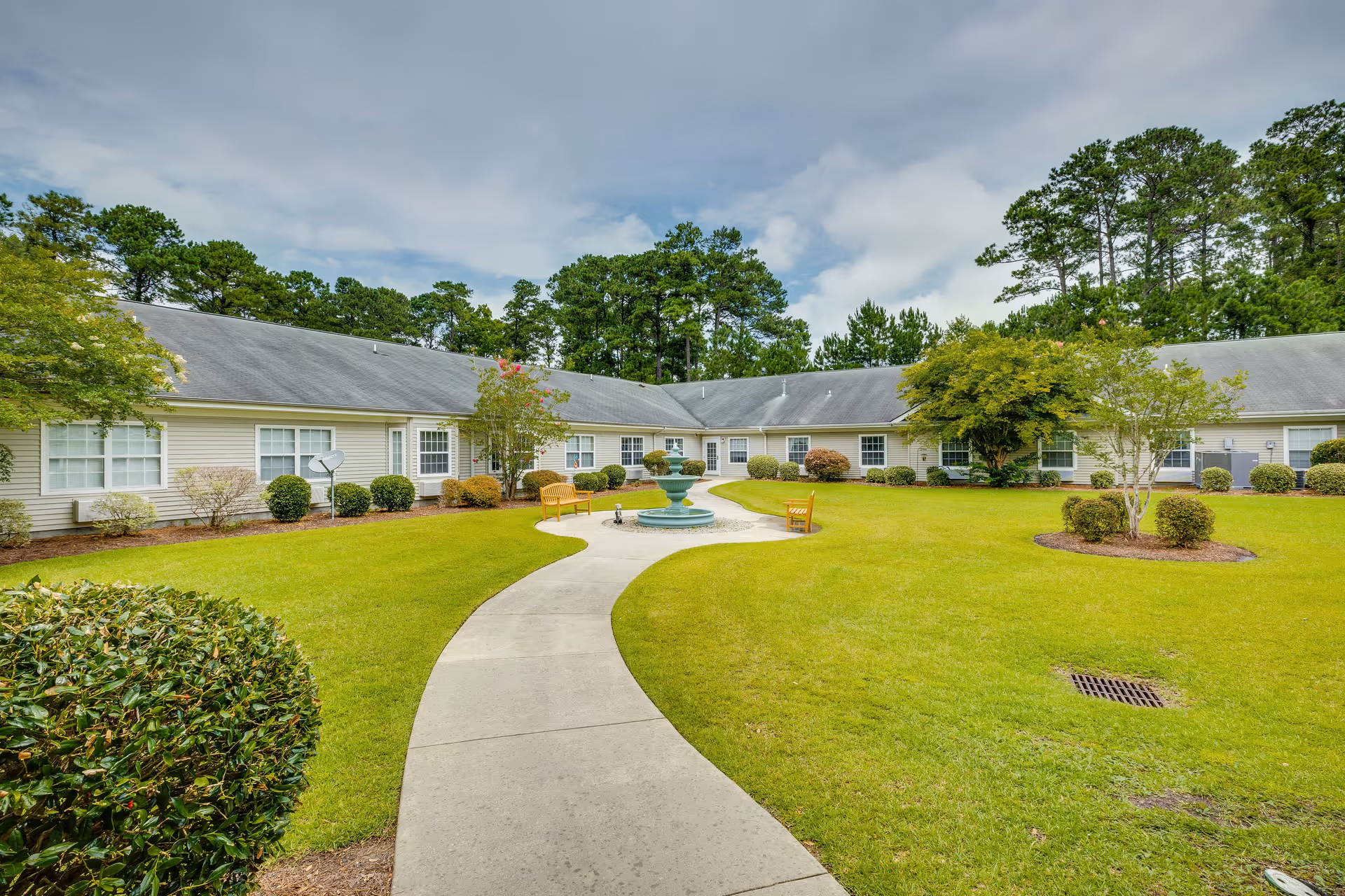 A well-maintained outdoor courtyard area at The Legacy of North Myrtle Beach featuring a curved concrete walkway leading to a central fountain surrounded by benches, green grass, bushes, and trees with a single-story building in the background under a partly cloudy sky.