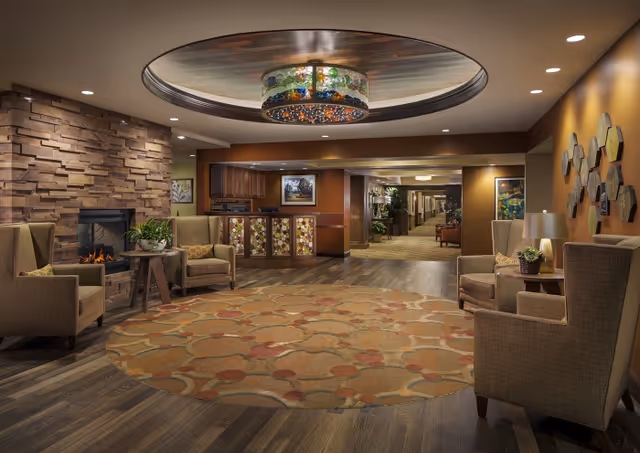 Warmly lit senior living lobby with armchairs around a patterned rug, a stone fireplace, decorative overhead light fixture, and a reception desk in the background.