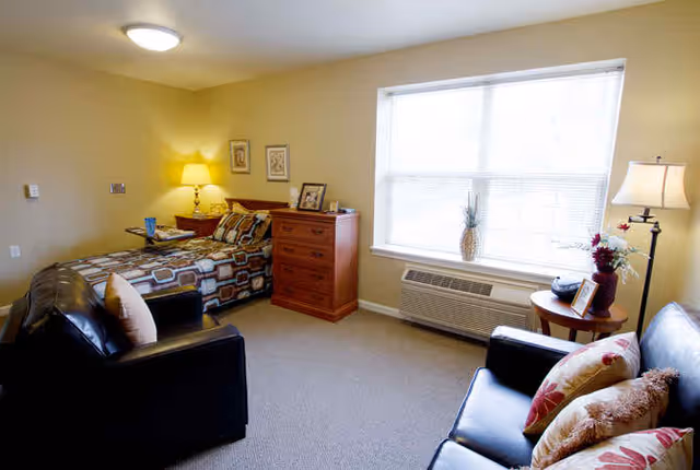A bright furnished studio bedroom with a bed, dresser, sofa, armchairs, side tables, and a large window.