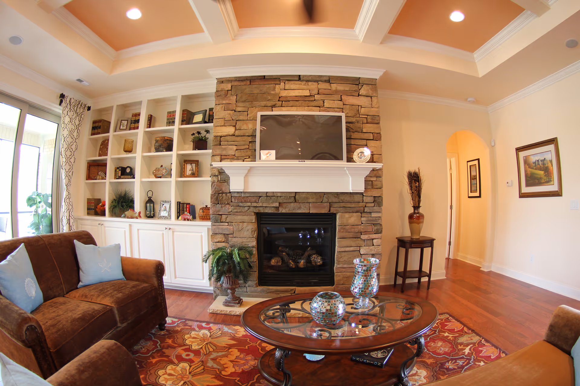 A cozy living room with a stone fireplace and a mounted flat-screen TV above it. The room features built-in white shelves filled with decorative items and books, a brown sofa with light blue pillows, a glass coffee table with decorative vases, a colorful patterned rug, hardwood floors, and a ceiling with recessed lighting and crown molding. There is an arched doorway leading to another room and framed artwork on the walls.