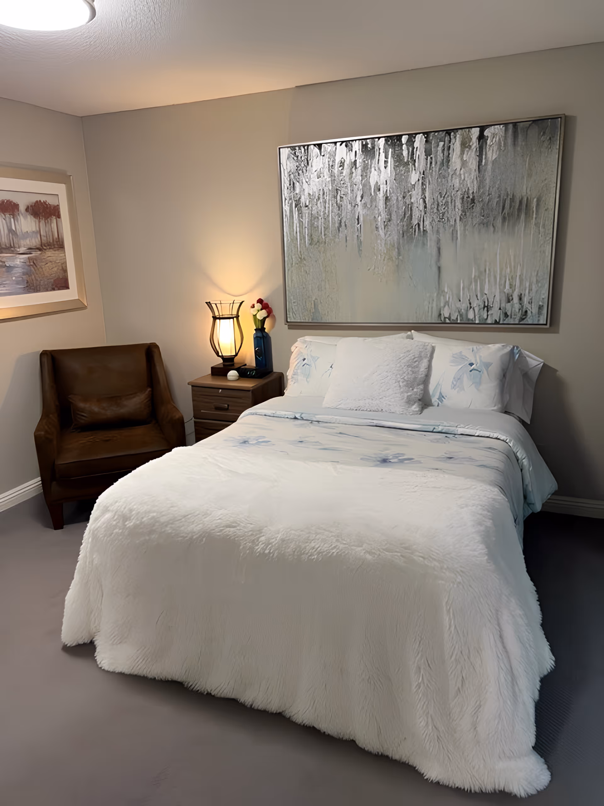 A cozy bedroom with a neatly made bed covered in a white fluffy blanket and floral sheets. Above the bed hangs a large abstract painting with silver and gray tones. To the left of the bed is a wooden nightstand with a decorative lamp and a vase with flowers. A brown leather armchair is positioned in the corner next to a framed landscape painting on the wall.