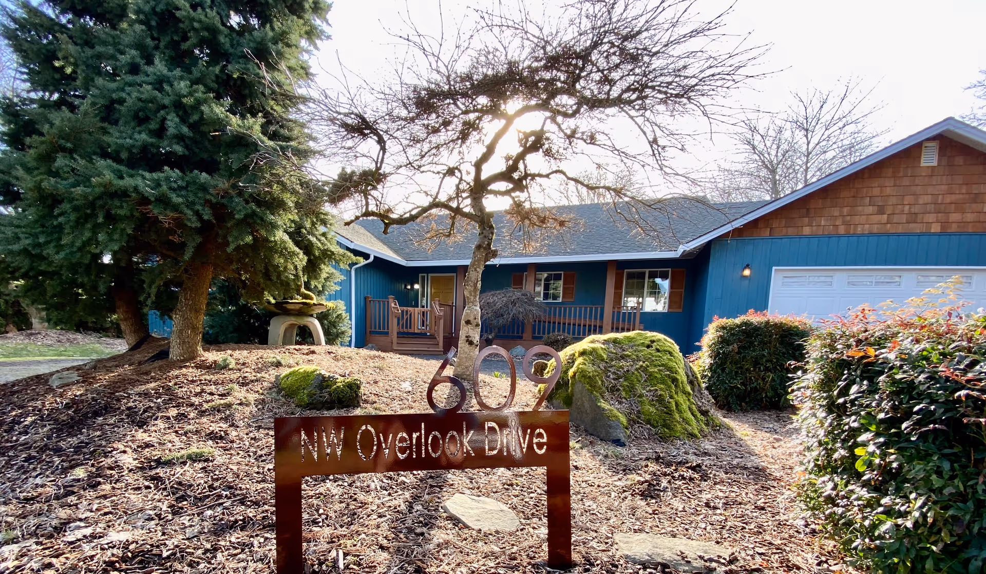 Front exterior view of a single-story house with blue siding and a wooden porch. The house has a garage with a white door on the right side. In front of the house, there is a landscaped area with mulch, bushes, a moss-covered rock, and a small tree. A metal address sign in the foreground reads '602 NW Overlook Drive'.