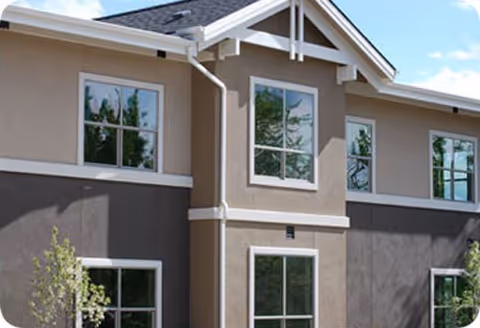 Exterior view of a two-story residential building with beige and brown walls, multiple windows, and a gable roof under a partly cloudy sky.