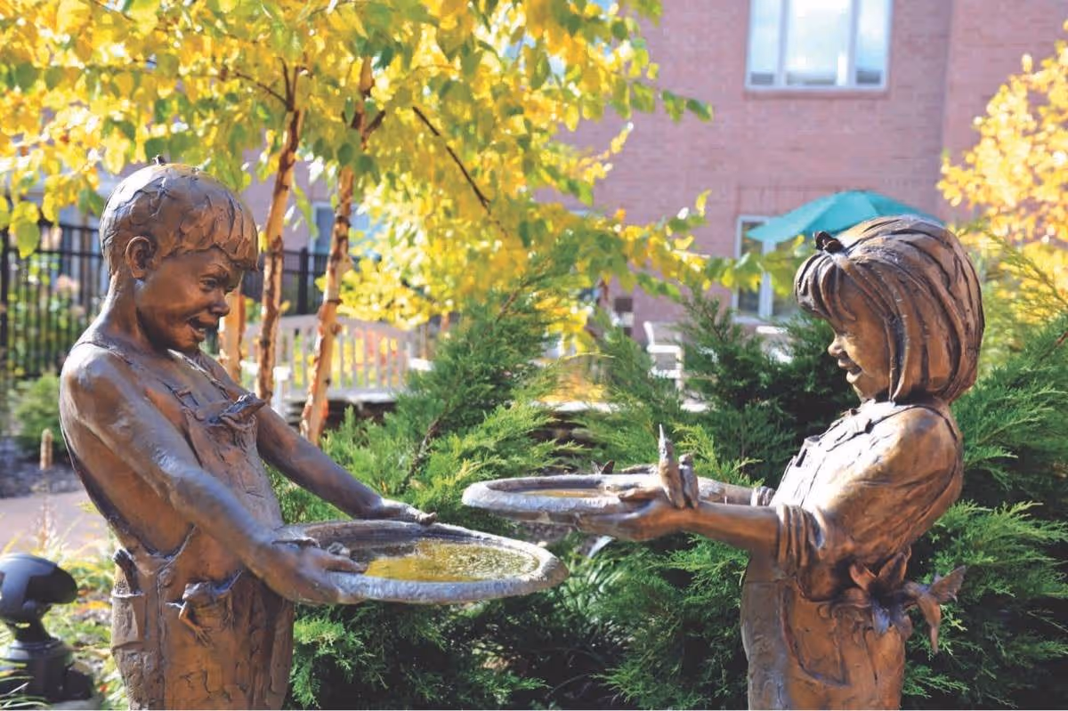 Bronze statues of a boy and a girl holding birdbaths, surrounded by greenery and trees with yellow leaves, with a brick building and patio furniture in the background.
