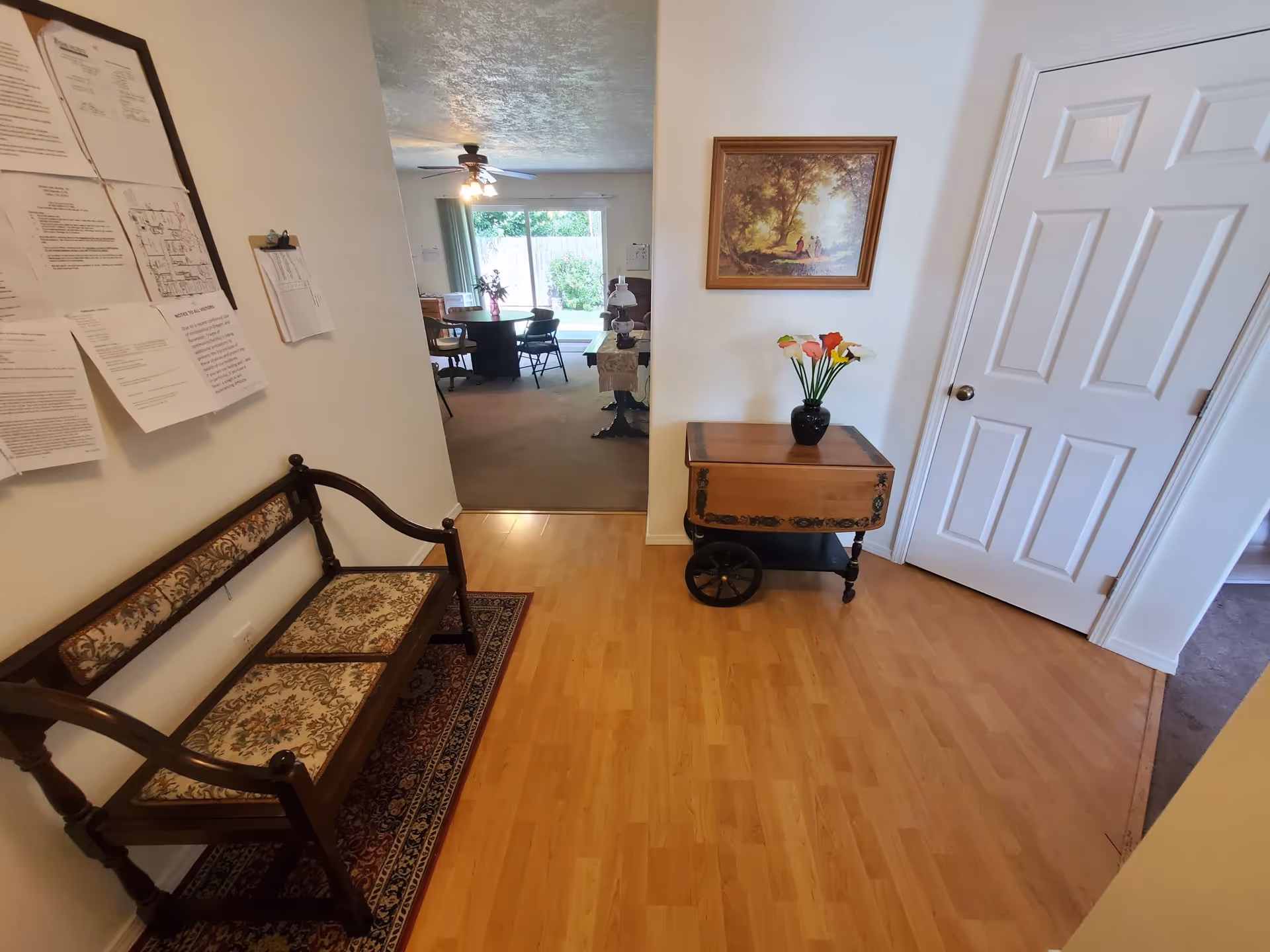 Interior view of a senior living facility hallway with wooden flooring, a vintage wooden bench with floral upholstery on the left, and a small wooden table with a vase of colorful flowers on the right. A framed painting hangs above the table. In the background, there is a dining area with a round table, chairs, a ceiling fan, and a large window with curtains letting in natural light.