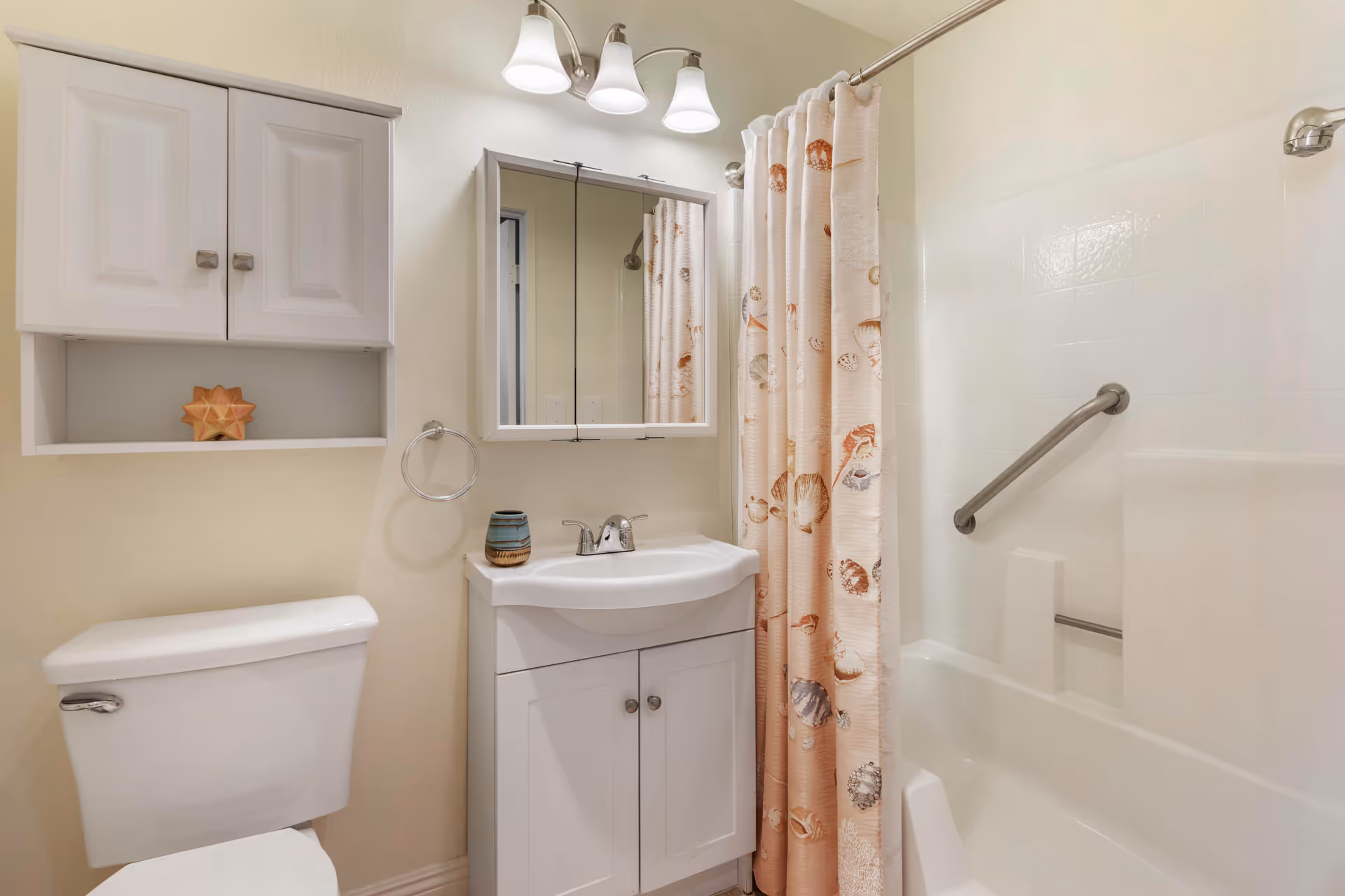A clean bathroom featuring a white toilet, a white vanity with a sink and a small decorative cup, a wall-mounted cabinet with two doors and an open shelf holding a star-shaped decoration, a mirror with three overhead lights, and a bathtub with a shower curtain decorated with seashell patterns and a grab bar.