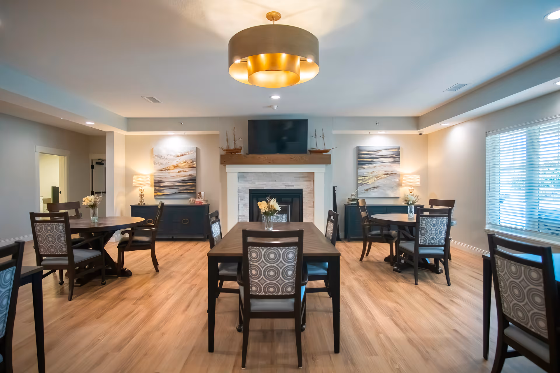 A bright and spacious dining room with wooden floors, multiple round and rectangular tables surrounded by chairs with patterned backs. The room features a fireplace with a TV mounted above it, flanked by two dark blue cabinets with lamps and decorative paintings on the walls. A large modern ceiling light fixture hangs in the center of the room, and a window with blinds allows natural light to enter.
