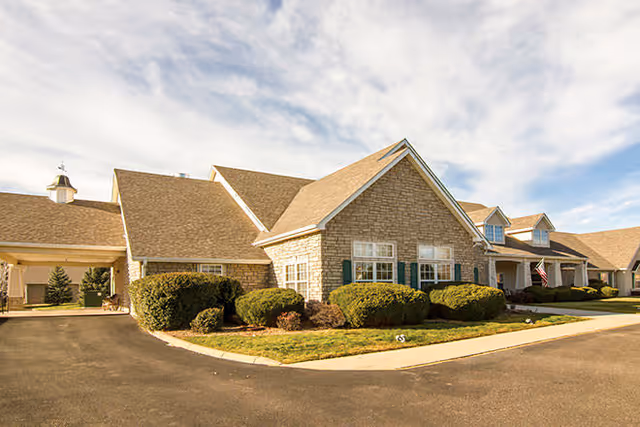 Front exterior of a single-story stone-clad senior living building with manicured shrubs, a driveway, and an American flag.