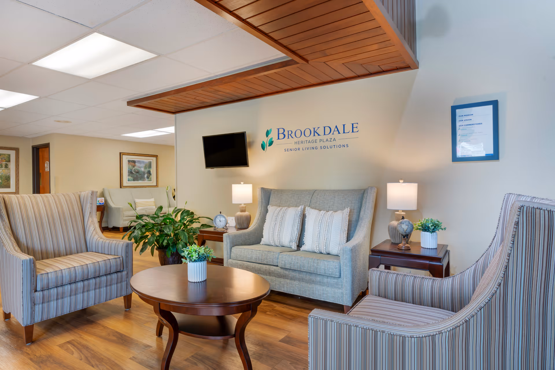 Comfortable senior living lounge with sofas, striped armchairs, a coffee table, plants, lamps, a wall-mounted TV and a 'Brookdale Heritage Plaza' logo on the wall.
