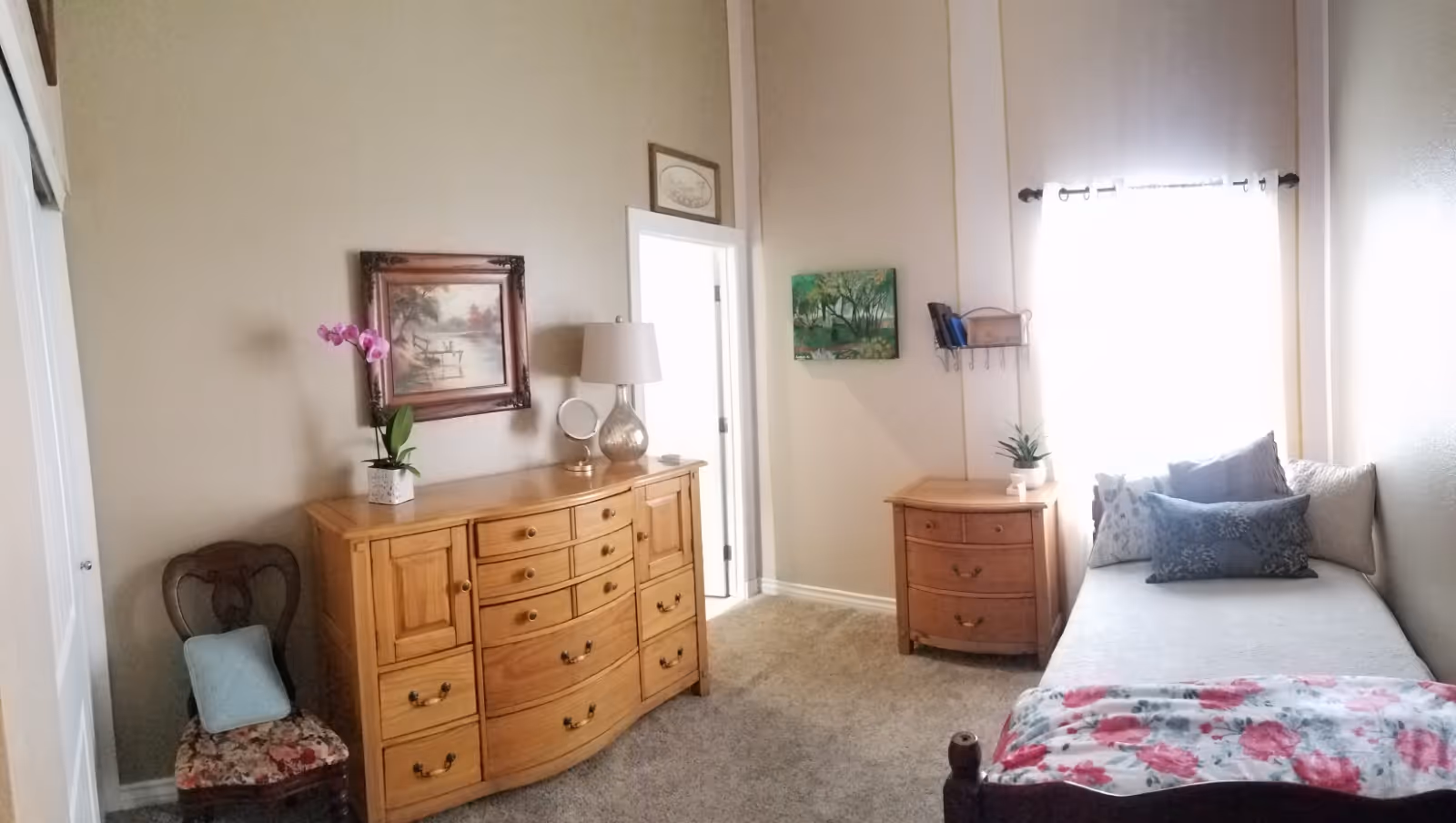 A cozy bedroom with beige walls and carpeted floor. There is a wooden dresser with multiple drawers, a table lamp, a small mirror, and a potted orchid on top. Next to the dresser is a wooden chair with a floral cushion and a blue pillow. On the right side, there is a daybed with several decorative pillows and a floral blanket. A small wooden nightstand with a plant and some books is beside the daybed. Two paintings hang on the walls, and a window with sheer curtains lets in bright natural light.