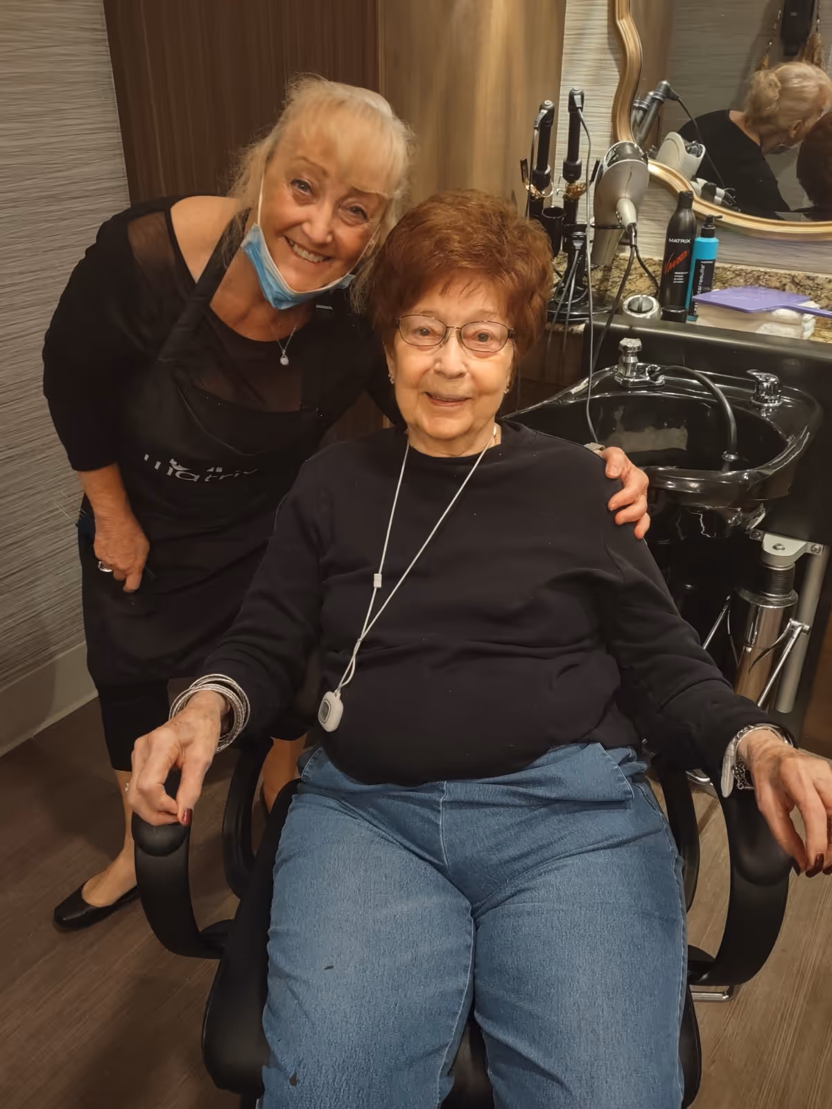 An elderly woman with glasses sitting in a salon chair, smiling, with a younger woman standing behind her wearing a black apron and a face mask pulled down under her chin. They are in a hair salon area with a black sink and hair styling tools visible in the background.