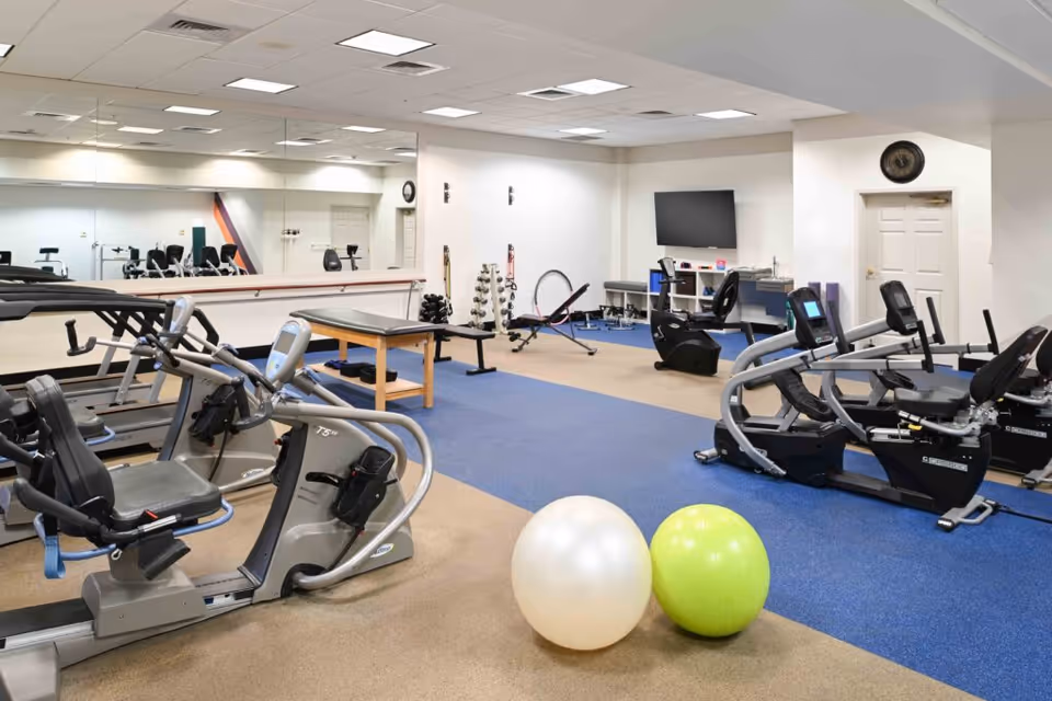 A fitness room with exercise equipment including recumbent bikes, treadmills, an exercise bench, dumbbells, two exercise balls, and a wall-mounted TV. The room has a blue and beige carpet, white walls, and a large mirror on one side.