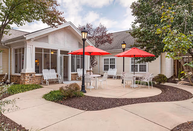 Outdoor patio area at Somerford House Frederick with white chairs and tables under red umbrellas, surrounded by a paved walkway, greenery, and a beige building with large windows and a door.