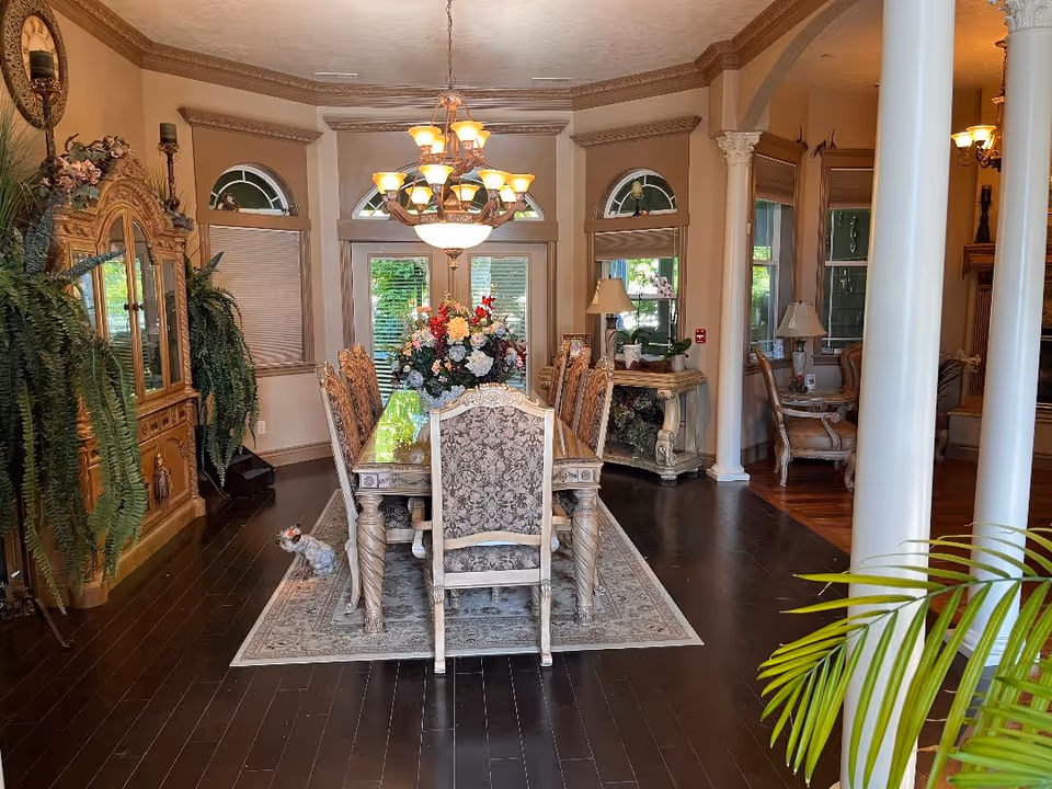 A spacious, elegant dining room with a long wooden dining table surrounded by ornate chairs. A large floral centerpiece decorates the table. The room features dark hardwood floors, a decorative area rug, a chandelier hanging above the table, and large windows with arched transoms. There are plants and decorative furniture pieces along the walls, and white columns partially frame the view into an adjacent sitting area.
