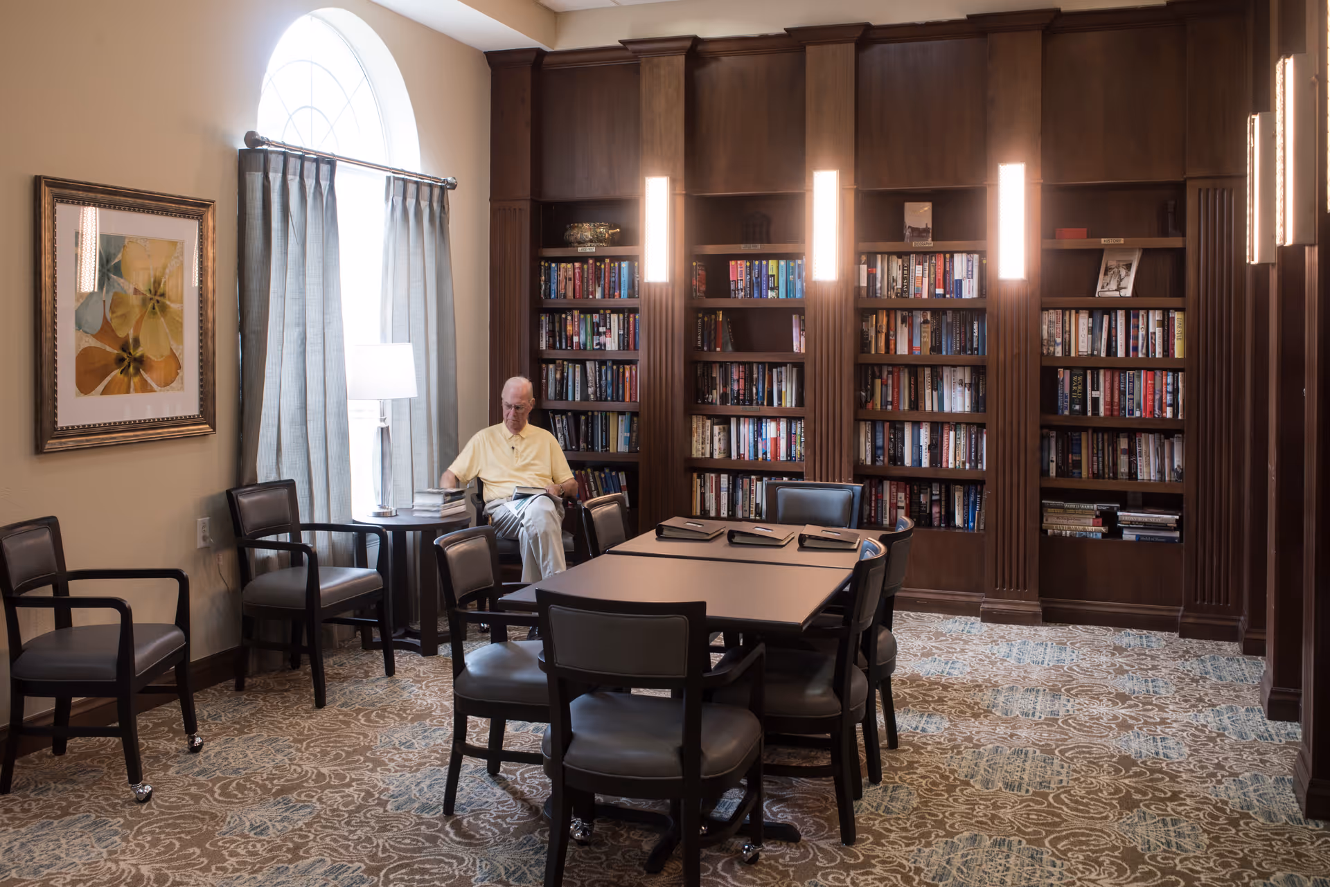A man reads in a library-style common room with built-in bookshelves, a table and several chairs.