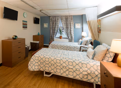 A bright shared bedroom with two twin beds, dressers, wall-mounted TVs and a window with patterned curtains.