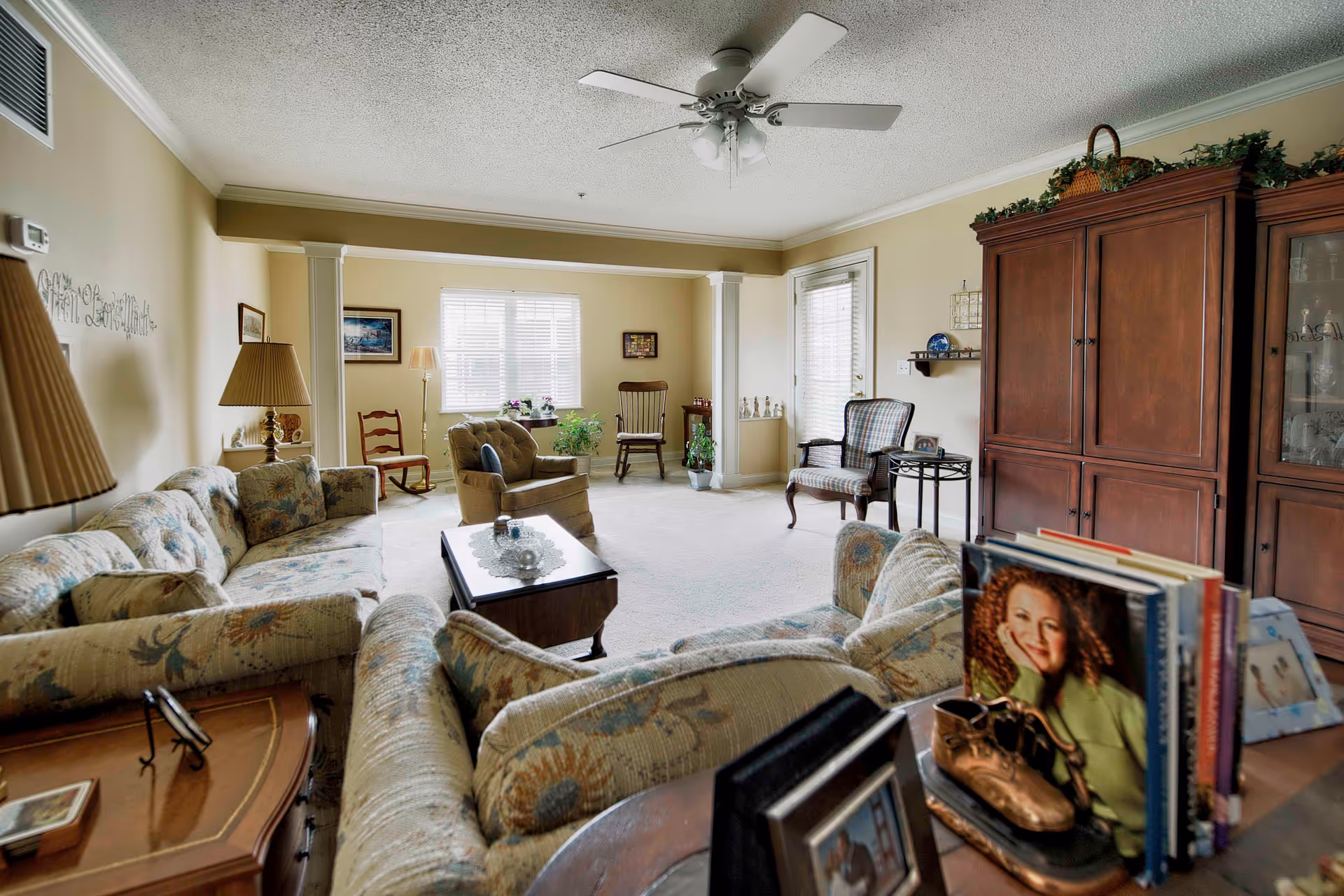 A cozy living room with floral patterned sofas, a wooden coffee table with a lace doily, several chairs near windows with blinds, a ceiling fan, and a large wooden cabinet. There are various decorative items including framed pictures, plants, and books on a side table.