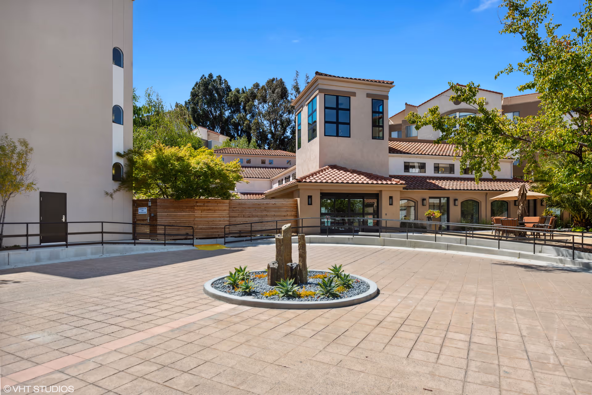 Outdoor courtyard area of a senior living facility with a circular stone planter featuring a small water fountain and succulents in the center. Surrounding the courtyard are beige buildings with red tile roofs, large windows, and some outdoor seating with tables and umbrellas. Trees and greenery are visible around the courtyard under a clear blue sky.