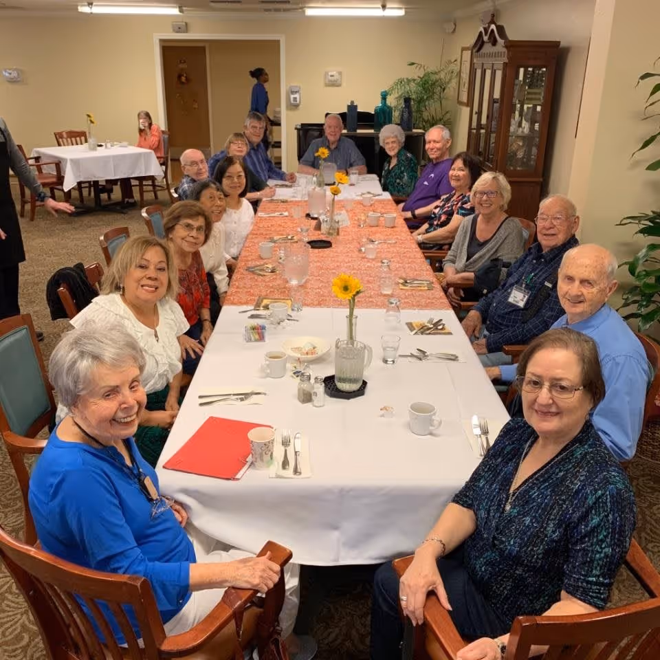 A group of elderly people sitting around a long dining table covered with white and orange tablecloths, smiling and posing for the photo in a dining room setting. The table has cups, utensils, pitchers of water, and small vases with yellow flowers. There are chairs and other tables in the background, along with a cabinet and some plants.