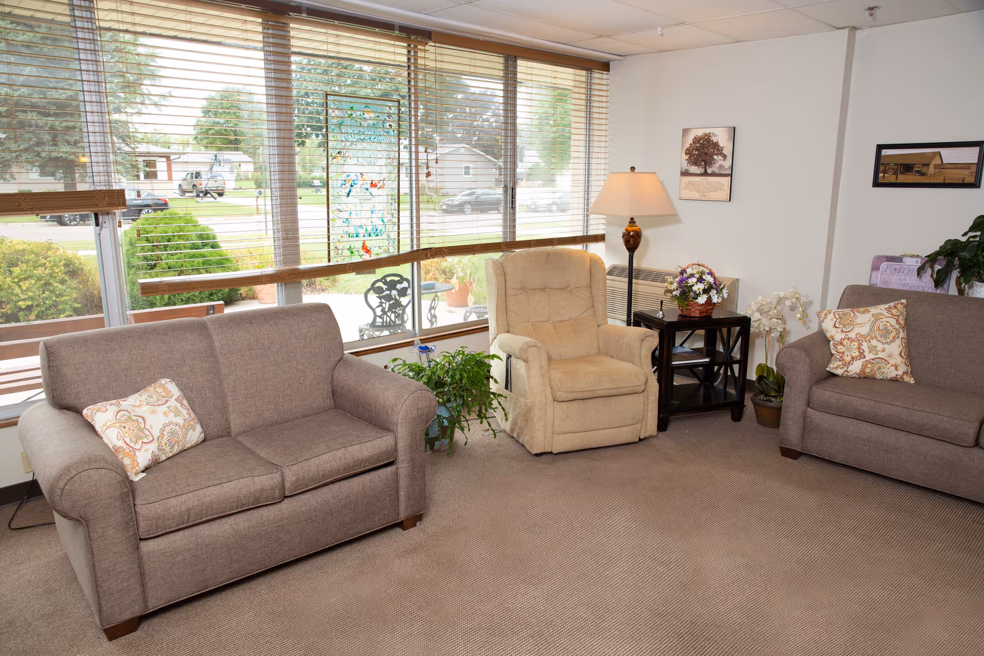 A cozy living room area with two brown sofas, each with a patterned throw pillow, and a beige recliner chair. There is a small black side table with a lamp and a basket of flowers between the recliner and one sofa. Large windows with blinds let in natural light and show a view of greenery and parked cars outside. The walls have framed pictures and there are several potted plants around the room.