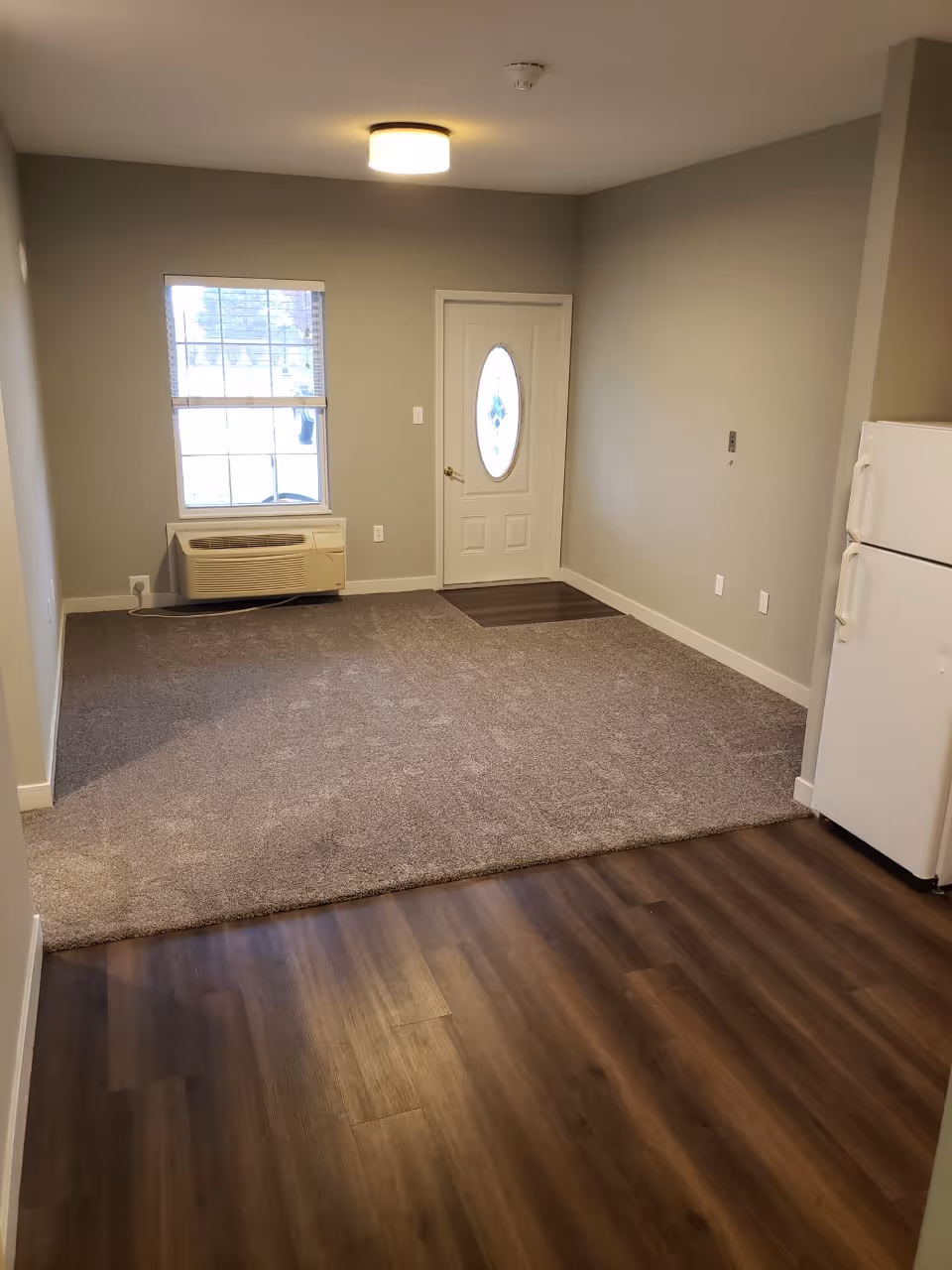 Empty interior room with a window, a white door with an oval glass panel, a beige carpeted area, and dark wood flooring. There is a white refrigerator on the right side and a wall-mounted air conditioning unit below the window.